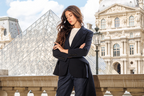 A woman posing in a black tailored blazer and matching trousers with black heels, set against the iconic Louvre Museum background in Paris.
